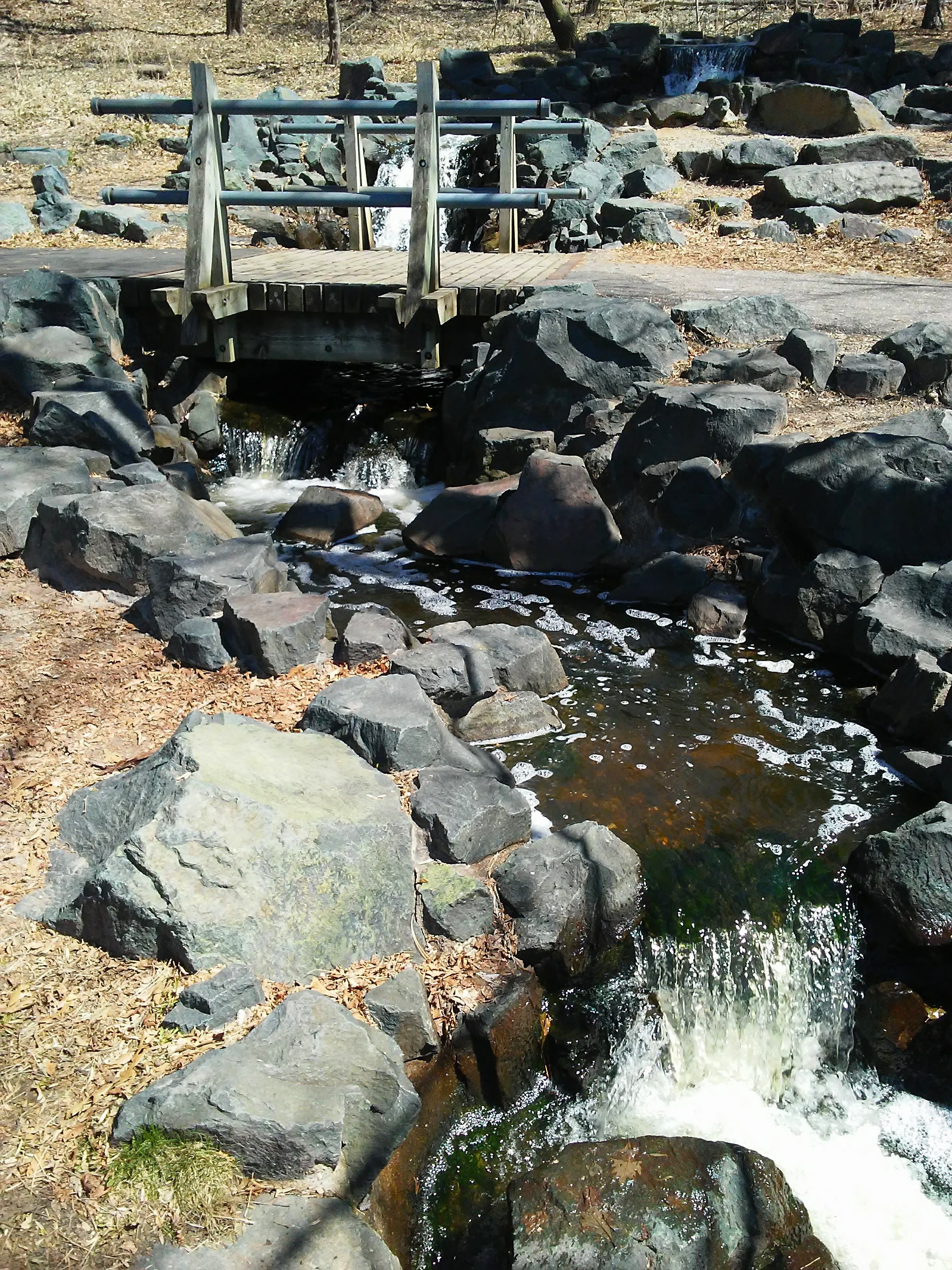 Bridge over a man-made creek with rocks and small waterfall.